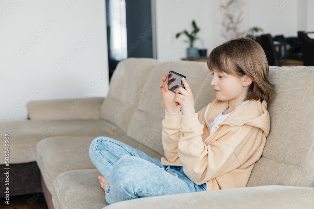 Schoolgirl is sitting on the sofa and playing mobile phone or chatting with friends. Children use technology. Modern gadgets and teenagers. Schoolgirl is sitting on the sofa and playing mobile phone or chatting with friends. Children use technology. Modern gadgets and teenagers.