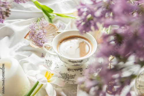 Spring composition with tulips and lilac flowers, candles and cup of coffee on white bed sheets. Cozy home atmosphere.