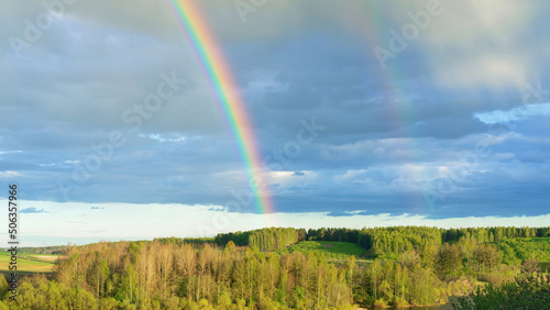 Double rainbow as a rare natural phenomenon against the backdrop of a hilly rural landscape. Bright real rainbow over the forest in early spring. The concept of weather forecast and climate change.