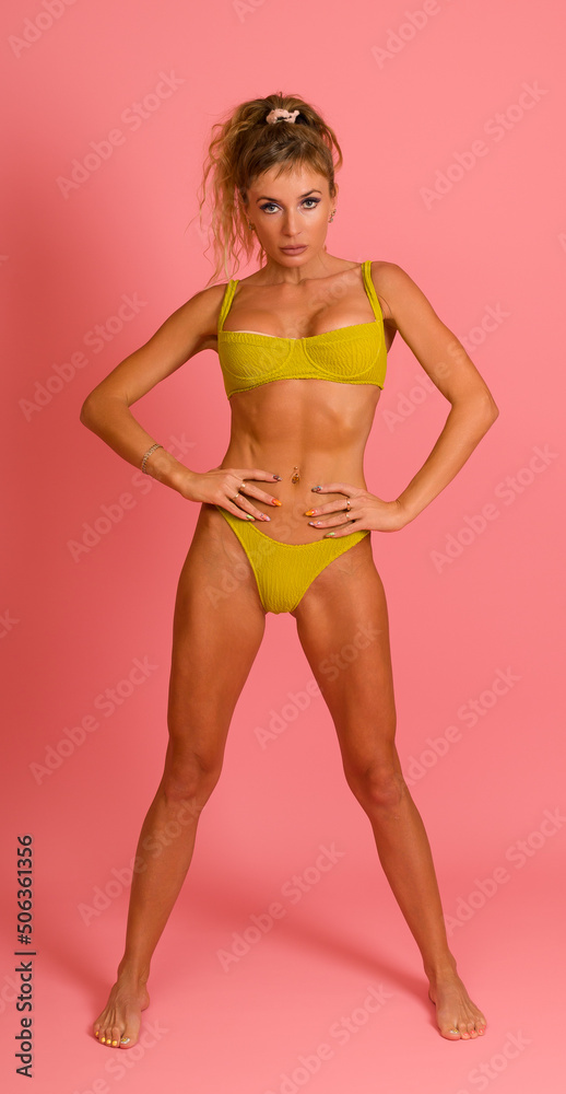 woman with a fit body in a yellow swimsuit posing while standing with her hands on her hips on a pink background.