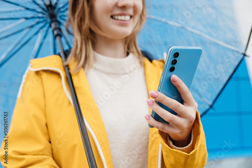 young caucasian woman dressed in a yellow raincoat holds a transparent umbrella and uses a smartphone, close-up