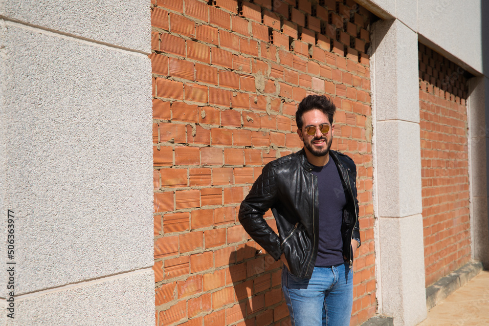 middle-aged man, handsome and dark, bearded and with a sculpted body on a brick wall. Man wearing black leather jacket, mirrored sunglasses, and jeans. Man poses for photo. Men's fashion concept.