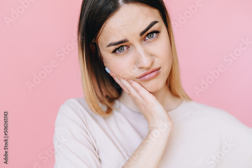 Portrait of a girl suffering from toothache frowns her face and looks at camera on pink background