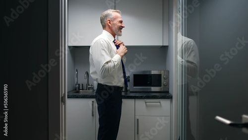 Senior businessman look into mirror and adjust tie standing in small office kitchen