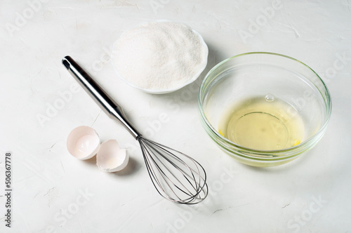 ingredients for the French meringue dessert - egg white, sugar, whisk and eggshell on a gray surface