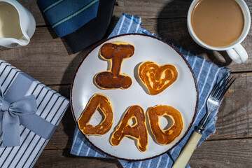 Father day pancakes. Homemade kids cooked father day pancakes with lettering I love dad, on plate, with coffee latte cup, gift box and necktie, wooden background top view copy space
