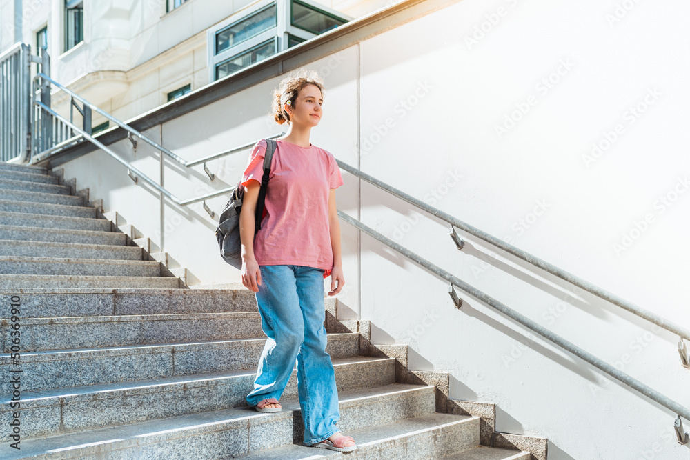 teenager girl, student of gymnasium, with backpack, steps down stairs in rays of sun. Positive emotions, young teenager girl, happy youth. Goes to meet happiness, airport building, university steps