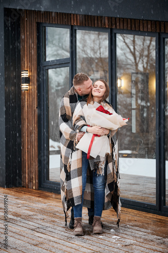 Full length of man wrapped in blanket kissing girlfriend in cheek while woman holding bouquet of flowers. Happy romantic couple standing outside scandinavian house barnhouse under winter snow.