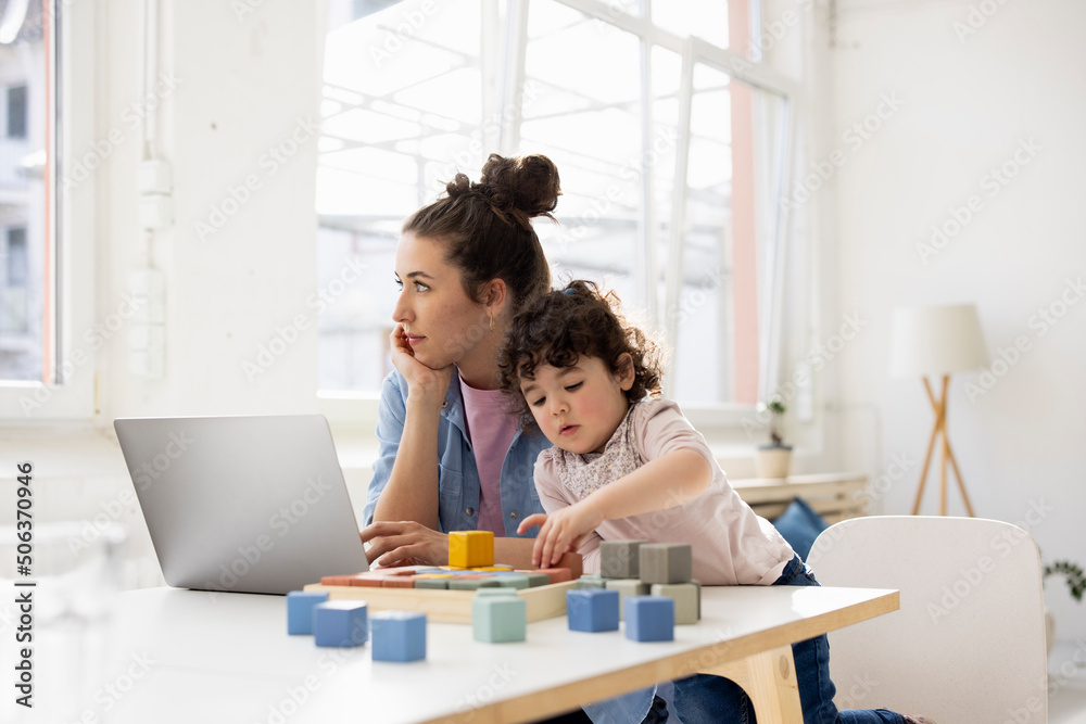 Mother working from home using laptop while daughter is playing with buiilding blocks Mother working from home using laptop while daughter is playing with buiilding blocks
