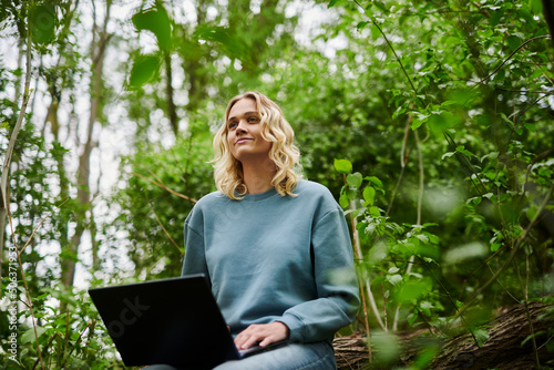 Contemplative freelancer sitting on fallen tree with laptop in forest photo