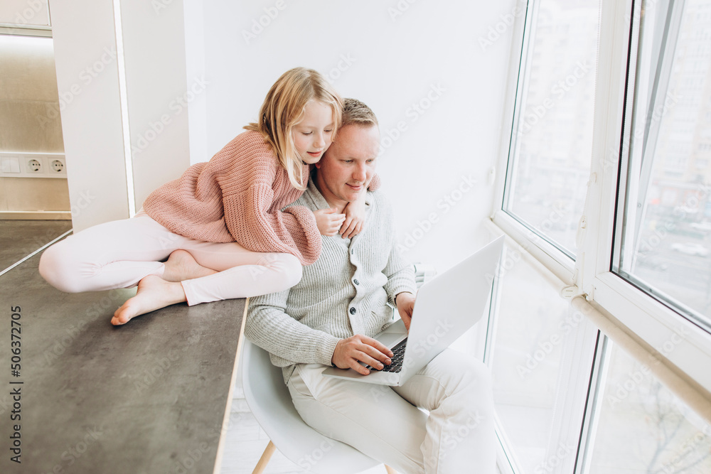 Daughter with father working on laptop sitting by window at home Daughter with father working on laptop sitting by window at home