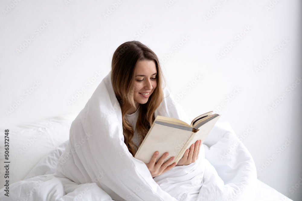 Smiling young woman wrapped in white blanket reading book on bed at home Smiling young woman wrapped in white blanket reading book on bed at home