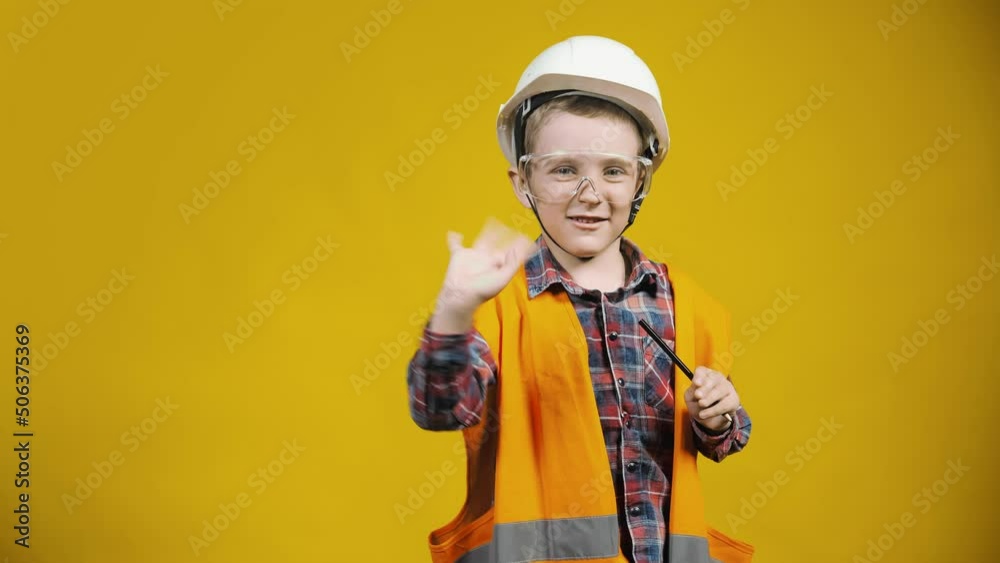 Portrait of a little construction boy in a helmet, waving and smiling at the camera on a yellow background. Children as adults Portrait of a little construction boy in a helmet, waving and smiling at the camera on a yellow background. Children as adults