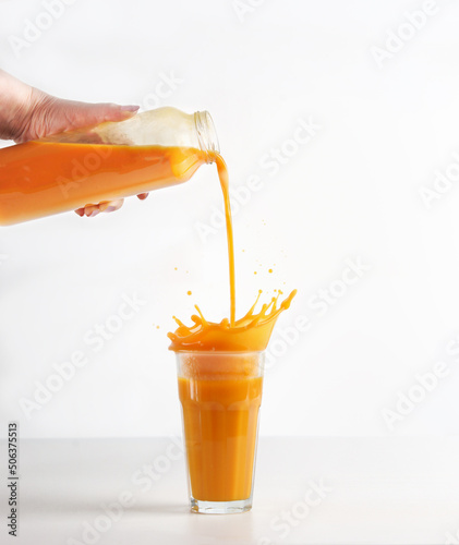 Women hand pouring homemade yellow juice from bottle in glass with splashing liquid at white background. Prepared healthy smoothie drink. Front view.