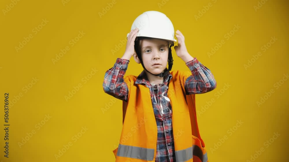 Portrait of a little boy-builder putting a helmet on his head on a yellow background. Children as adults Portrait of a little boy-builder putting a helmet on his head on a yellow background. Children as adults