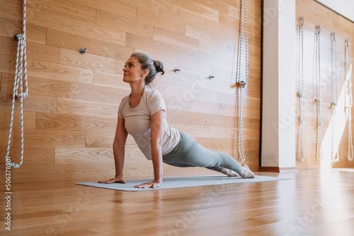 Full length portrait of attractive senior woman working out at home in living room, doing yoga or pilates exercise on grey mat, standing in plank pose (phalankasana)