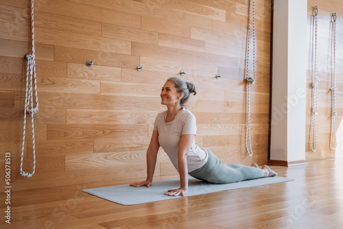 Full length view of the senior fit woman making cobra pose on yoga mat, exercising at home at the light room. Sport and recreation concept