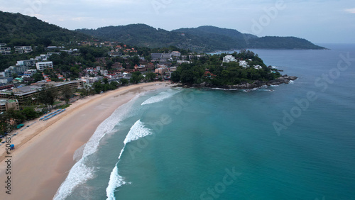 Early dawn on the island of Phuket. Top view from the drone on the sand and waves. People are walking along the beach. Calm environment. Green hills and clouds in sky. Lights of the city are shining © SergeyPanikhin