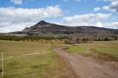 Mountain landscape, peak rises above the forest, mountain against the blue sky, clouds float, country road stretches across the field, journey trekking in nature