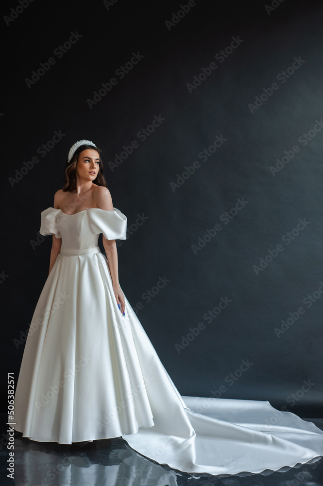 Beautiful young sexy model female bride with dark hair in gorgeous glossy long light wedding dress posing in dark studio Beautiful young sexy model female bride with dark hair in gorgeous glossy long light wedding dress posing in dark studio