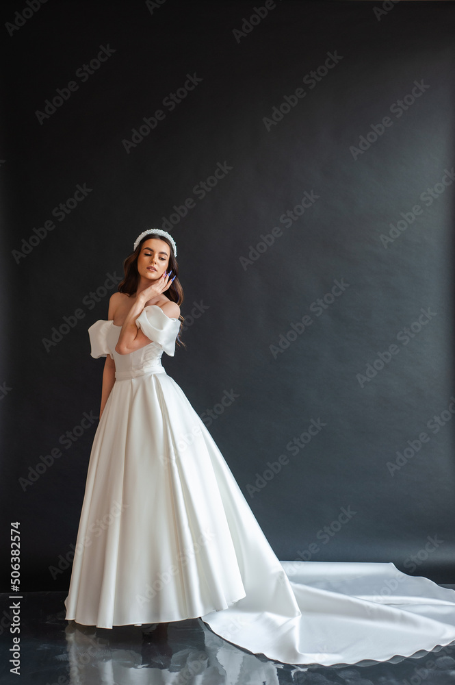 Beautiful young sexy model female bride with dark hair in gorgeous glossy long light wedding dress posing in dark studio Beautiful young sexy model female bride with dark hair in gorgeous glossy long light wedding dress posing in dark studio