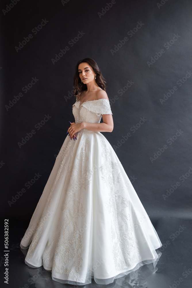 Beautiful young sexy model female bride with dark hair in gorgeous glossy long light wedding dress posing in dark studio