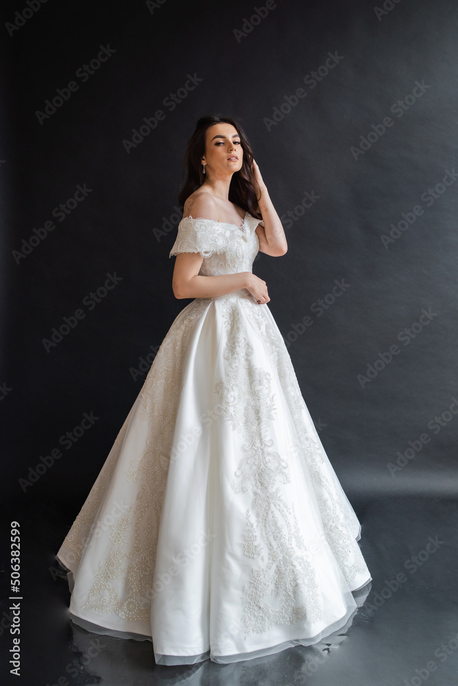 Beautiful young sexy model female bride with dark hair in gorgeous glossy long light wedding dress posing in dark studio