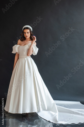 Beautiful young sexy model female bride with dark hair in gorgeous glossy long light wedding dress posing in dark studio