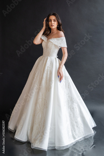 Beautiful young sexy model female bride with dark hair in gorgeous glossy long light wedding dress posing in dark studio