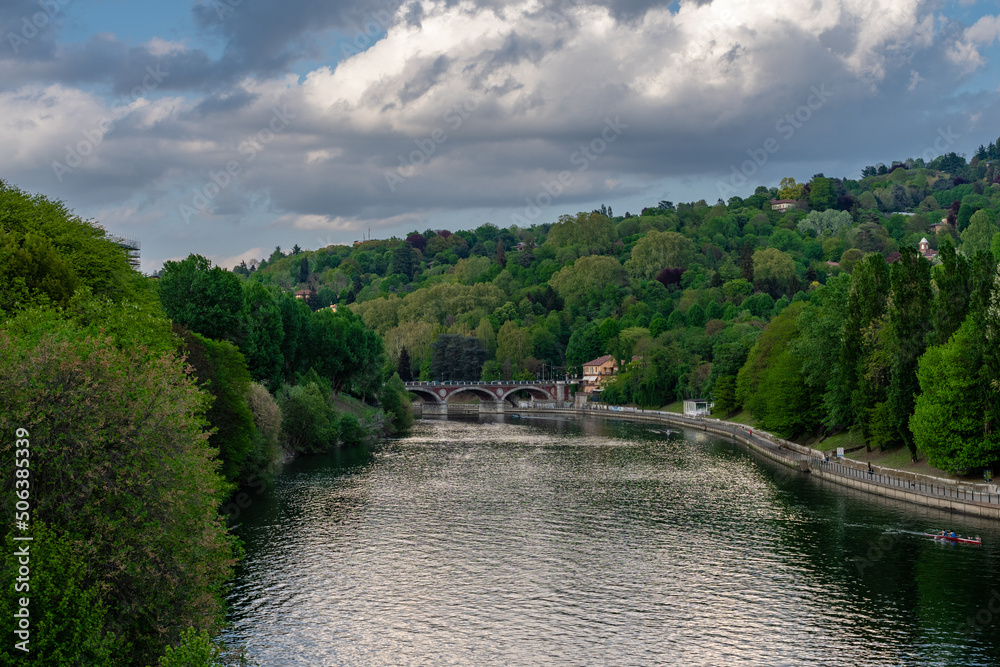 Fototapeta premium Natural landscape of the river Po in Torino on a spring afternoon. Green and leafy trees. Cloudy sky. And an old bridge that crosses the river.