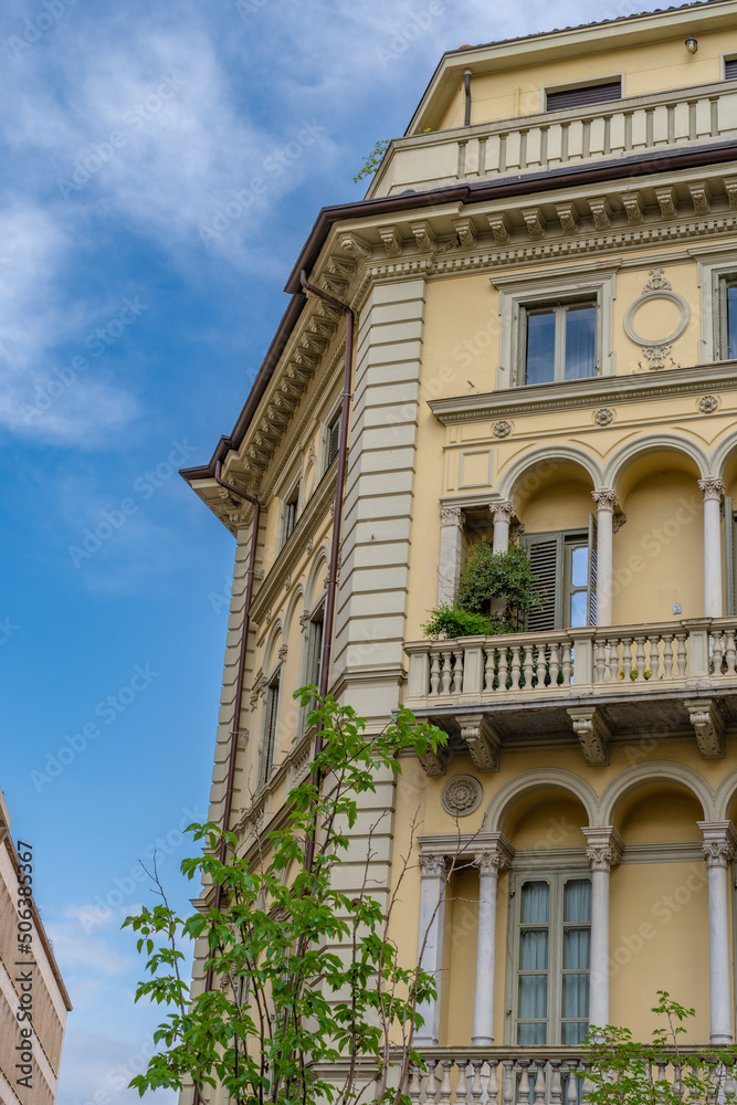 Facade of old, European building, with balconies with plants and arches. Behind a blue sky.