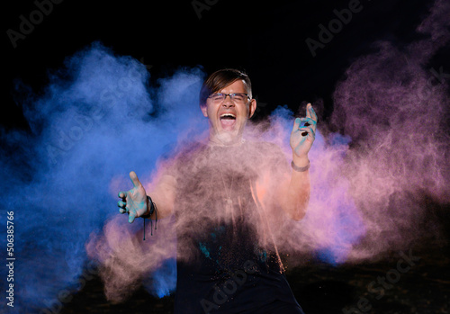 A young caucasian emotionally guy is standing strewn with holi paints on a dark background