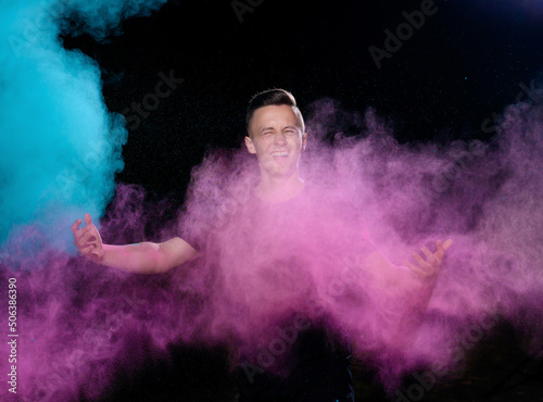 A young caucasian emotionally guy is standing strewn with holi paints on a dark background