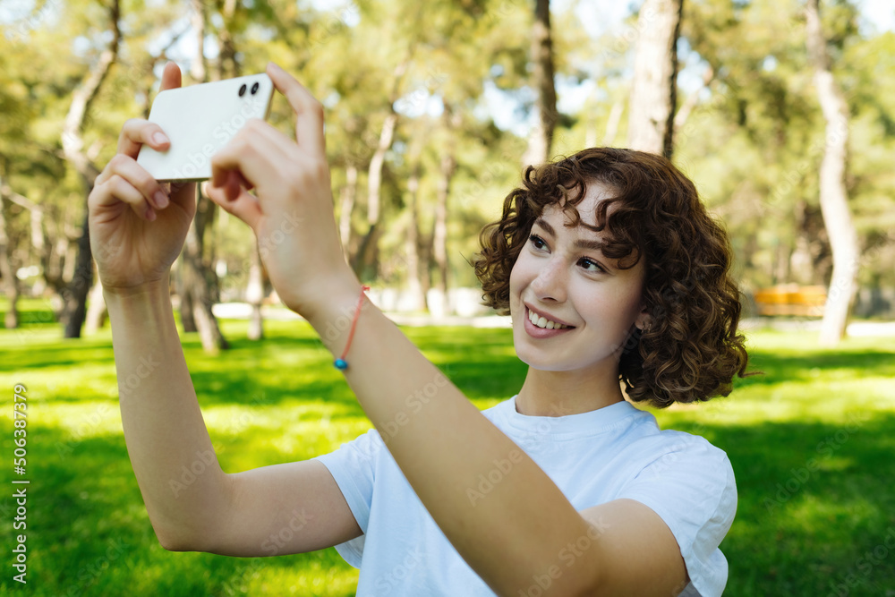 Redhead woman wearing white tee taking horizontal photo with her phone on green city park, outdoors. Landscape photo, outdoor lifestyle concepts. Redhead woman wearing white tee taking horizontal photo with her phone on green city park, outdoors. Landscape photo, outdoor lifestyle concepts.