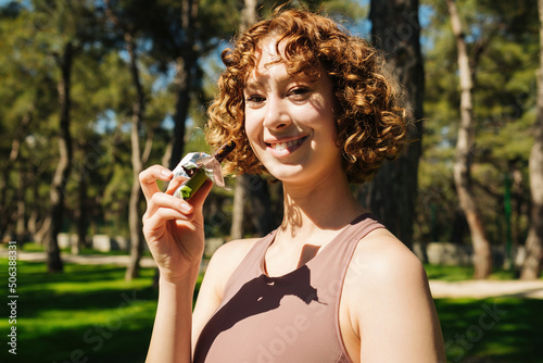 Portrait of young sporty redhead woman resting while biting a nutritive bar. Athletic woman eating a protein bar. Fitness beautiful woman eating a energy snack outdoor.