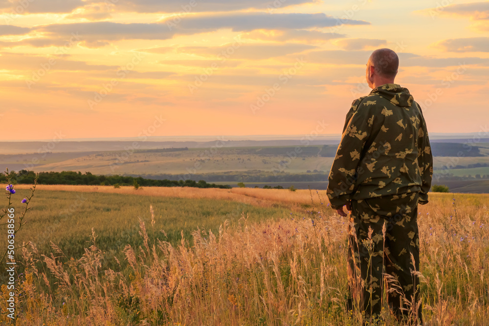 A European male farmer, 40-45 years old, stands in full growth in a field in the summer at dawn in the warm sun against the backdrop of the sunset sky and horizon. Man looking into the distance