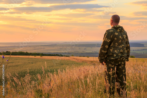 A European male farmer  40-45 years old  stands in full growth in a field in the summer at dawn in the warm sun against the backdrop of the sunset sky and horizon. Man looking into the distance