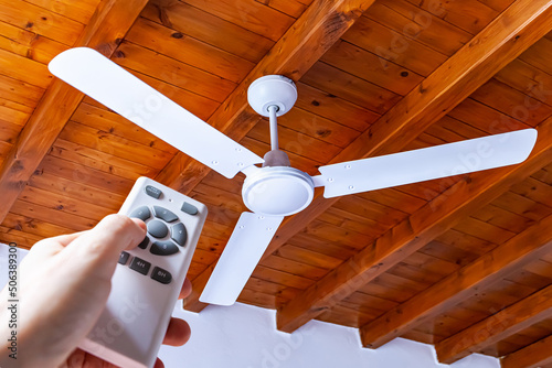 A man uses a remote control to turn on a white ceiling fan mounted in a house with wooden ceilings. photo