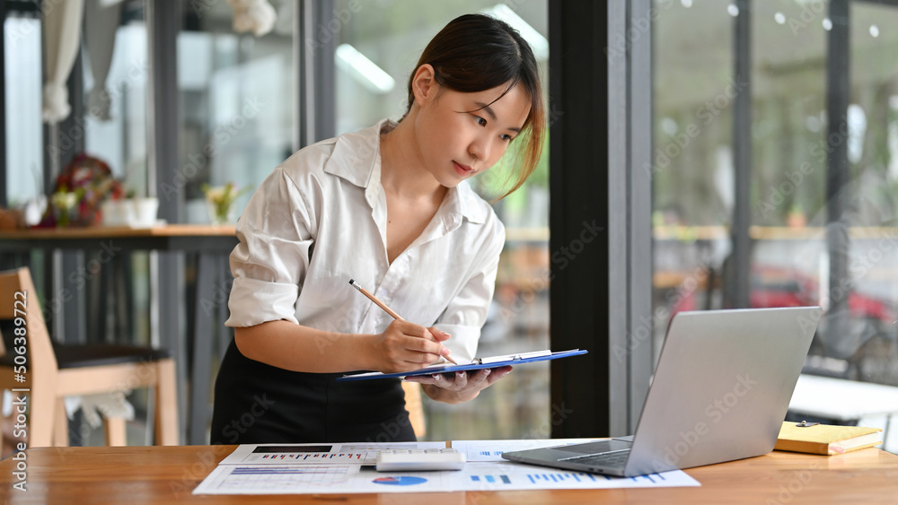 Female financial assistant looking the details on laptop screen and writing on clipboard Female financial assistant looking the details on laptop screen and writing on clipboard