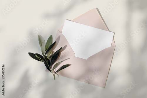 Summer Mediaterranean wedding stationery still life. Blank greeting card, imvitation mockup. Blush pink envelope in sunlight, beige table background in sunlight. Green olive tree branches. Flat lay photo