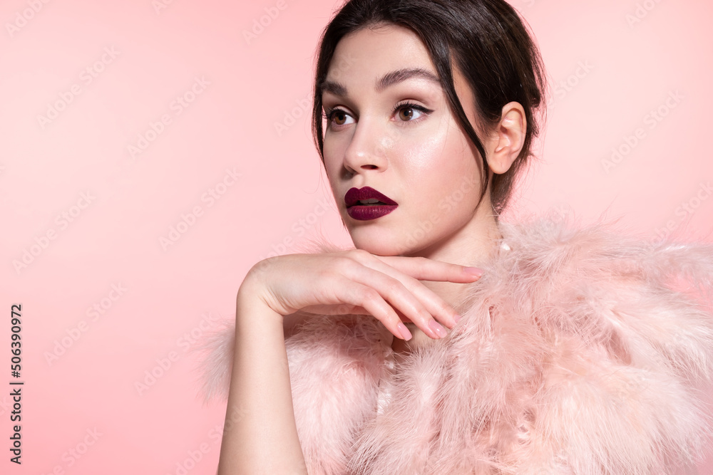 Fashion  girl with glowing healthy skin, perfect festive make up, celebratory attire posing on pink studio background. Fairy pretty delightful woman wearing holiday feathers boa looking aside.