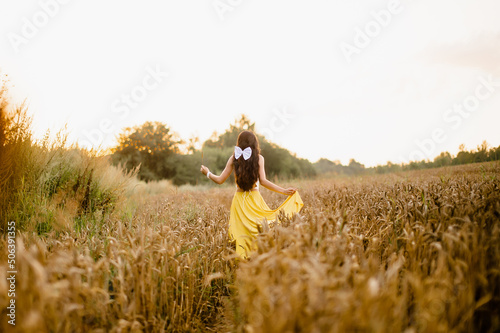 A girl with long hair and a white bow stands in a yellow skirt in a field with spikelets © Julia Jones