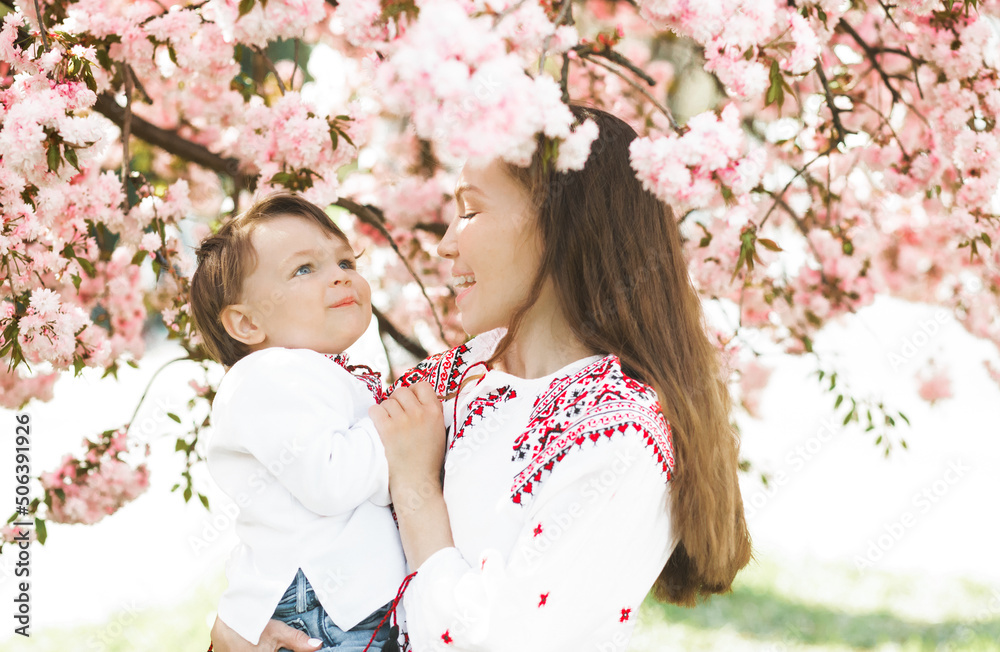 Fototapeta premium Little boy on hands of mother. Woman playing with child outside in blooming spring garden. Portrait of family of two people. Spring landscape background.