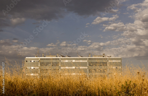 Low angle view of a modern building and a field of yellow wildflowers under a dramatic cloudy blue sky.