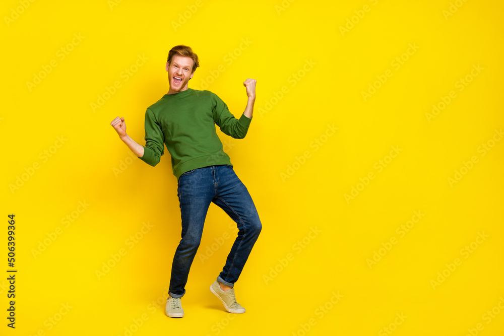 Full length photo of pretty lucky man wear green pullover rising fists empty space isolated yellow color background Full length photo of pretty lucky man wear green pullover rising fists empty space isolated yellow color background