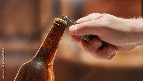Close-up man hand opens lid fresh light beer in sweating from cold dark brown glass bottle with lid opener on blurry background. Light lager is opened with help of bottle opener. photo