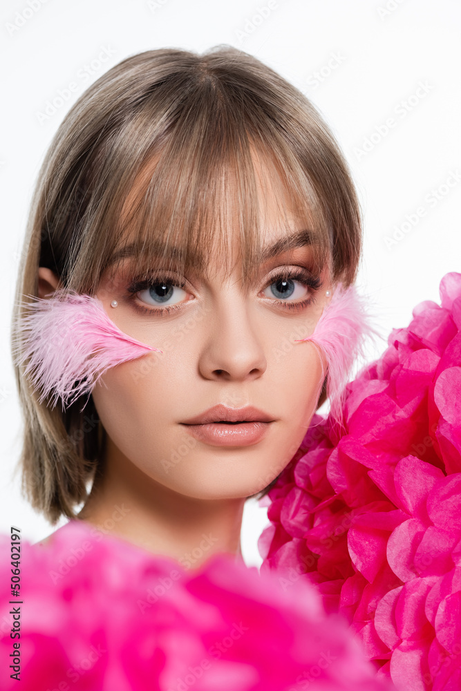 young woman with decorative elements in makeup, bangs and pink feathers on cheeks near flowers isolated on white young woman with decorative elements in makeup, bangs and pink feathers on cheeks near flowers isolated on white