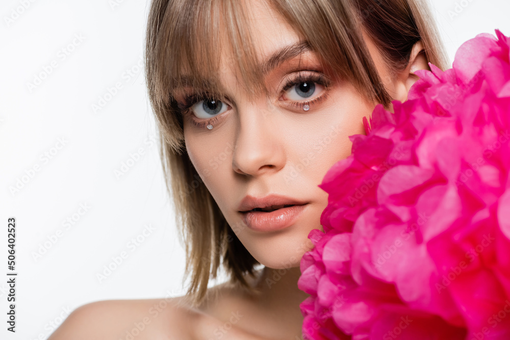 young woman with bangs and rhinestones under blue eyes near pink flower isolated on white
