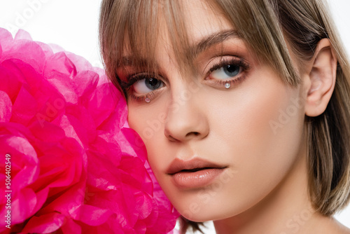 pretty young woman with bangs and shiny rhinestones under blue eyes near pink flower isolated on white