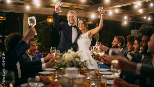 Beautiful Bride and Groom Celebrate Wedding at an Evening Reception Party. Newlyweds Propose a Toast to Happy Marriage  Standing at a Dinner Table with Best Multiethnic Diverse Friends.